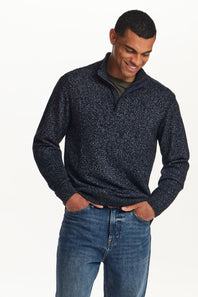A man wearing a dark navy and black textured quarter-zip sweater with blue jeans, standing against a plain white background.