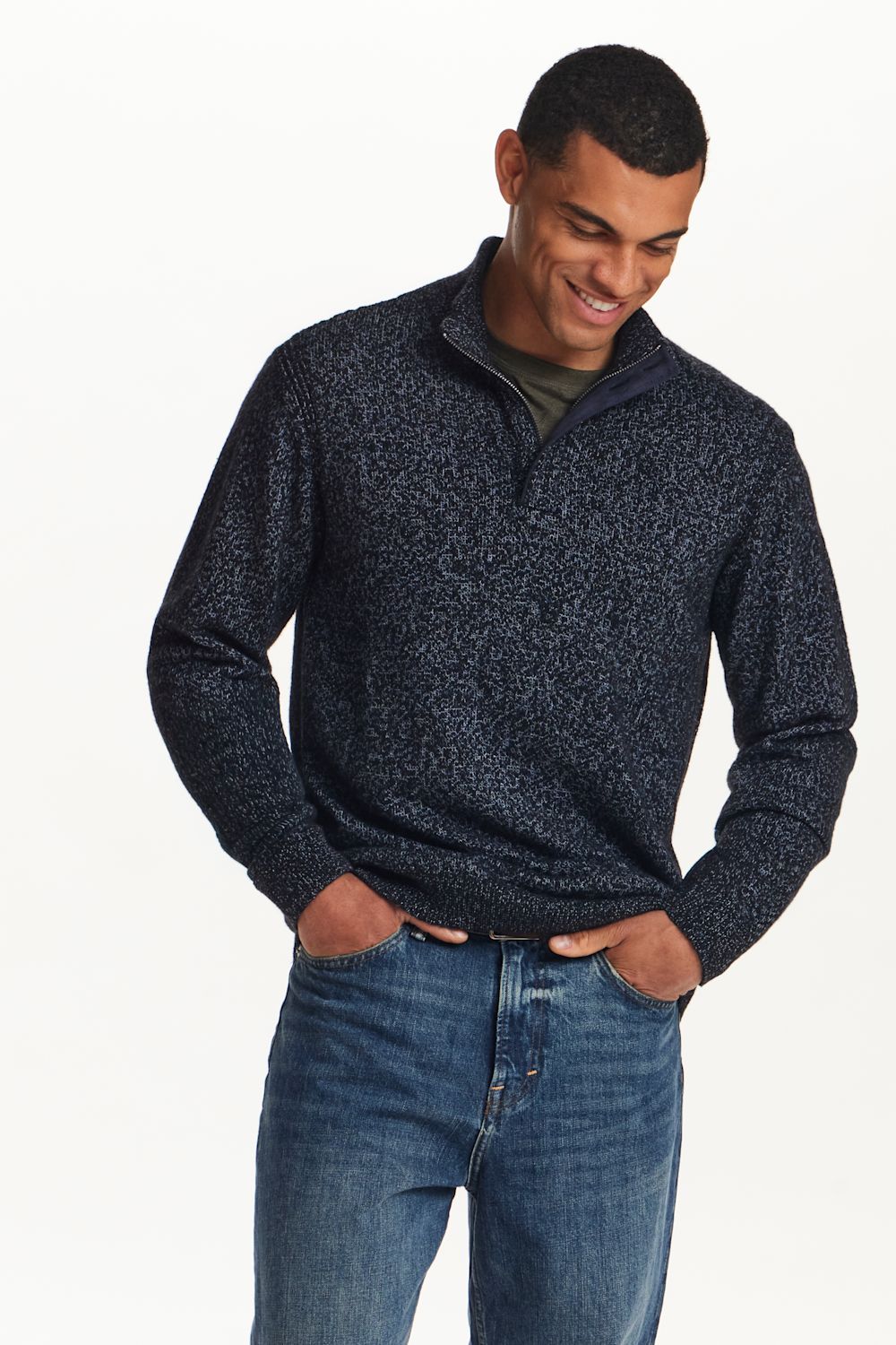 A man wearing a dark navy and black textured quarter-zip sweater with blue jeans, standing against a plain white background.
