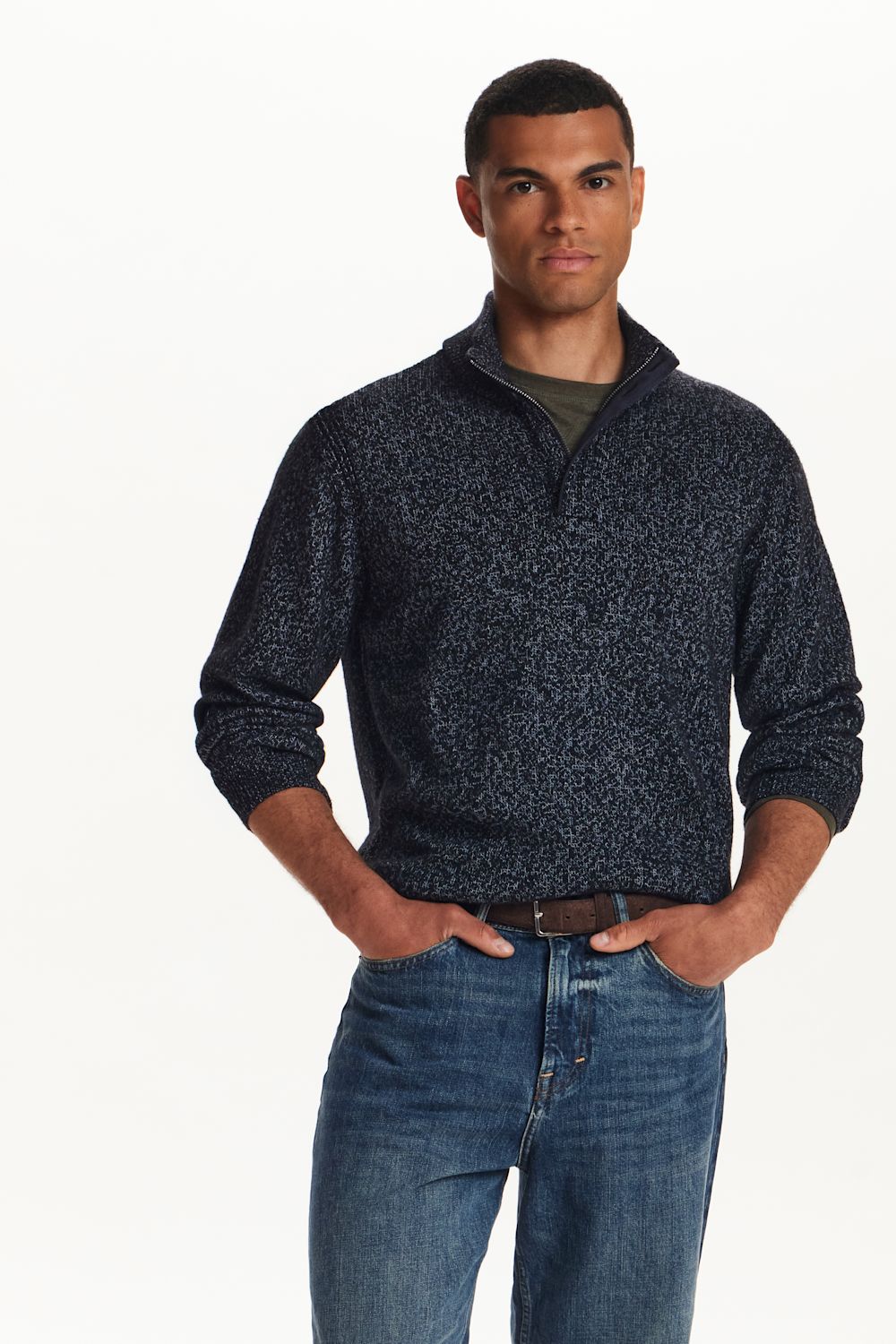 A man wearing a dark navy and black textured quarter-zip sweater over a green t-shirt and blue jeans, standing against a plain white background.