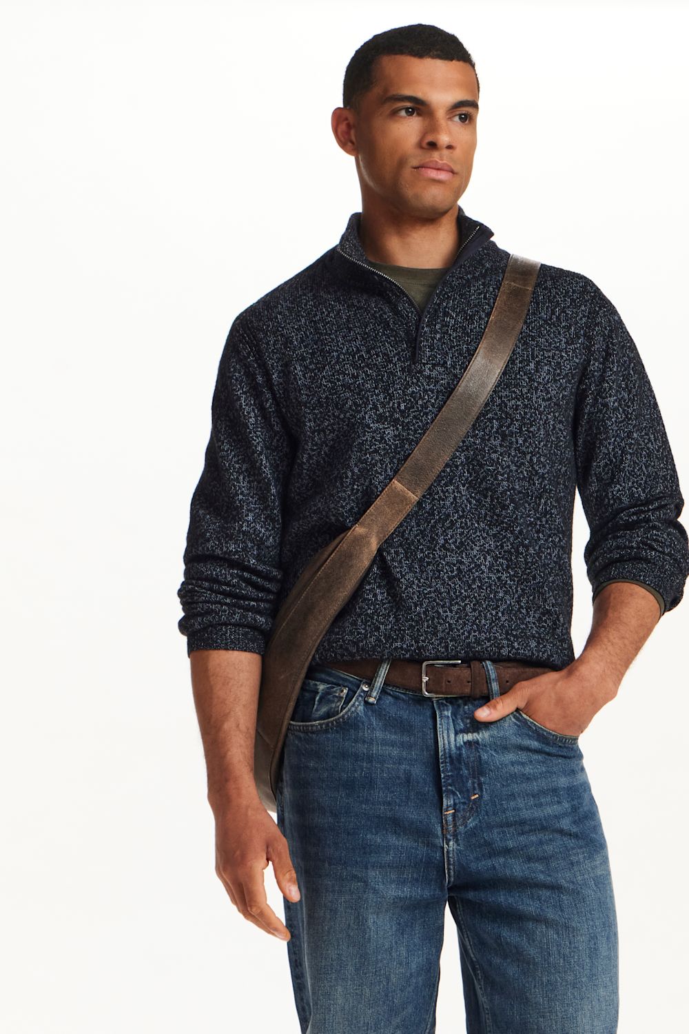 A man wearing a dark navy textured quarter-zip pullover sweater, blue jeans, and a brown leather crossbody bag, photographed against a clean white background.