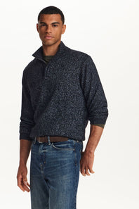 A man wearing a dark navy and black speckled quarter-zip sweater with blue jeans, standing against a plain white studio background.