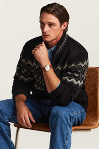 A man wearing a black knit cardigan with a gray and white geometric pattern, blue jeans, and a brown leather strap watch, sitting on a brown leather chair against a light background.