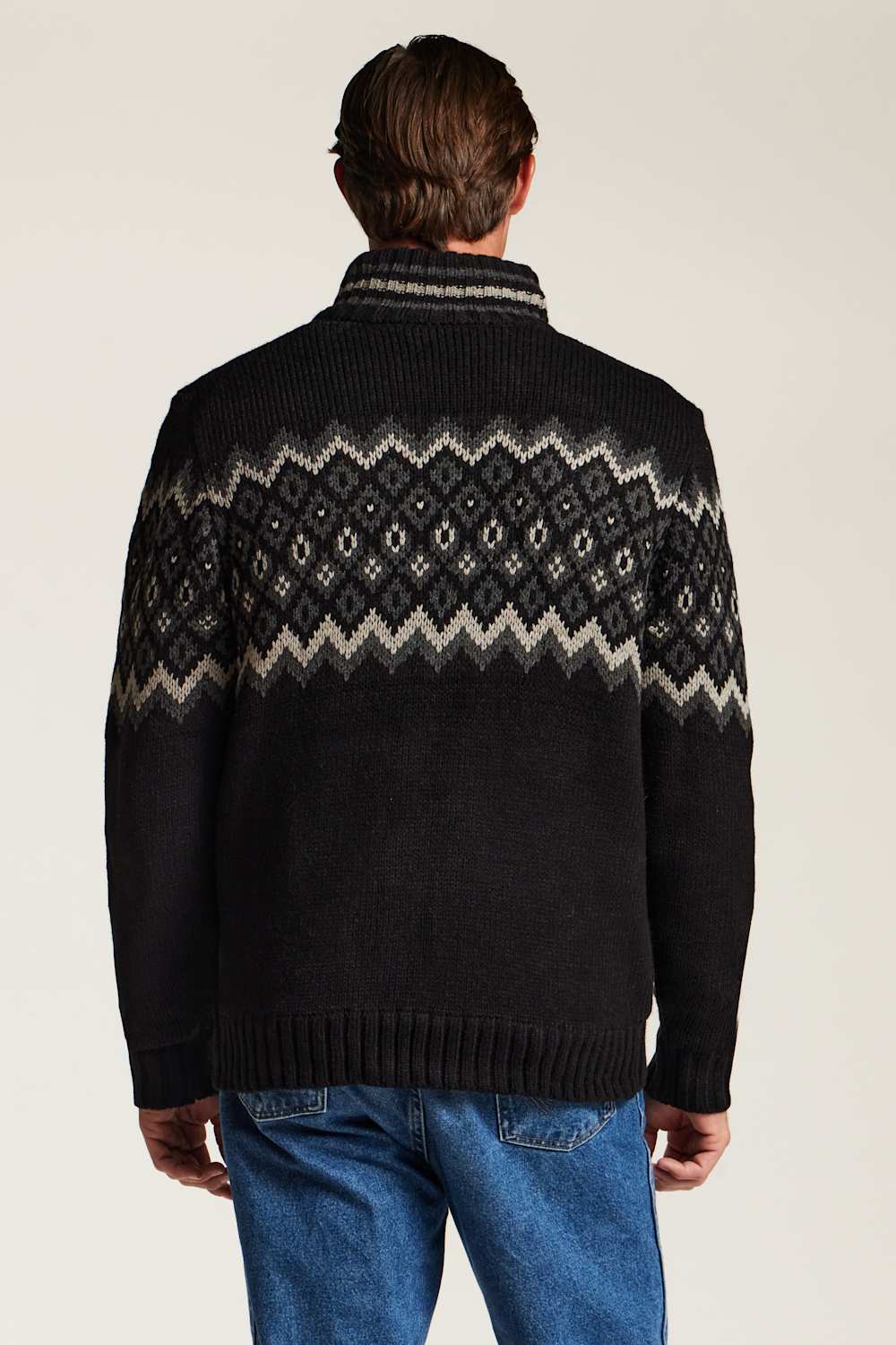 A man viewed from the back wearing a black knit sweater with a cream and gray geometric Fair Isle pattern across the shoulders and upper back, paired with blue jeans against a plain light background.