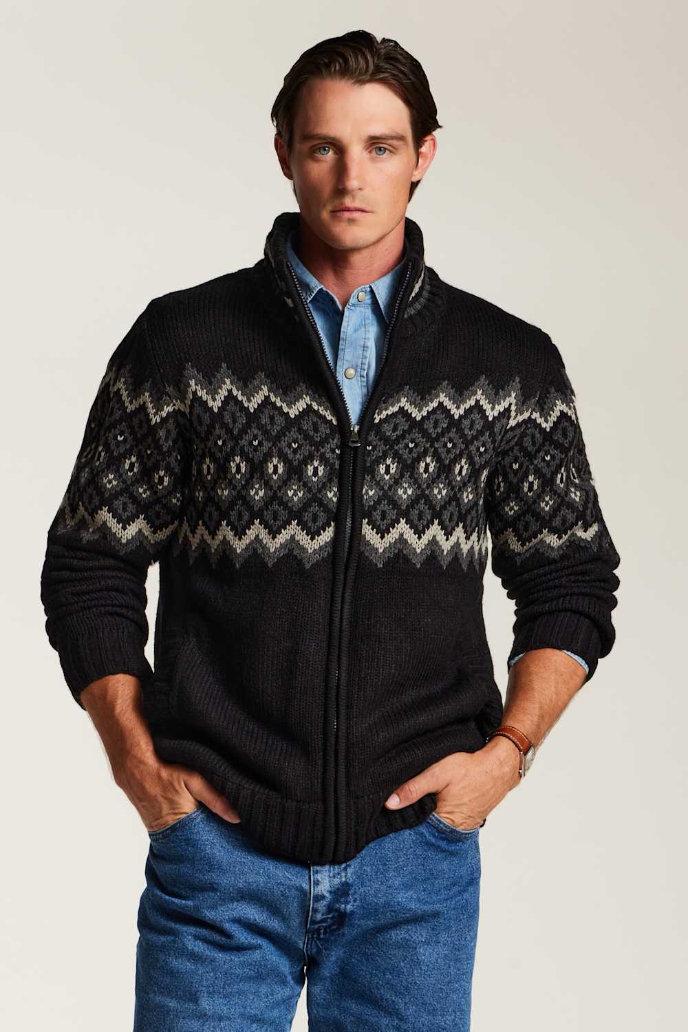 A man wearing a black zip-up knit sweater with a gray and white Fair Isle pattern across the chest and sleeves, layered over a light blue button-down shirt and blue jeans, against a plain light gray background.