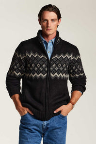 A man wearing a black zip-up knit sweater with a gray and white Fair Isle pattern across the chest and sleeves, layered over a light blue button-down shirt and blue jeans, against a plain light gray background.