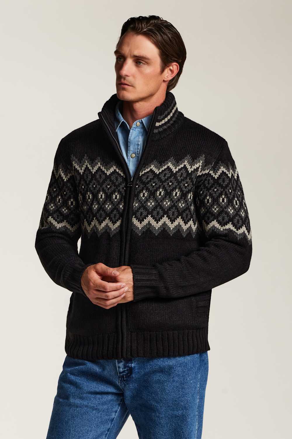 A man wearing a black knit zip-up sweater with a beige and gray geometric Fair Isle pattern across the chest and shoulders, layered over a light blue button-down shirt and blue jeans, against a plain light gray studio background.