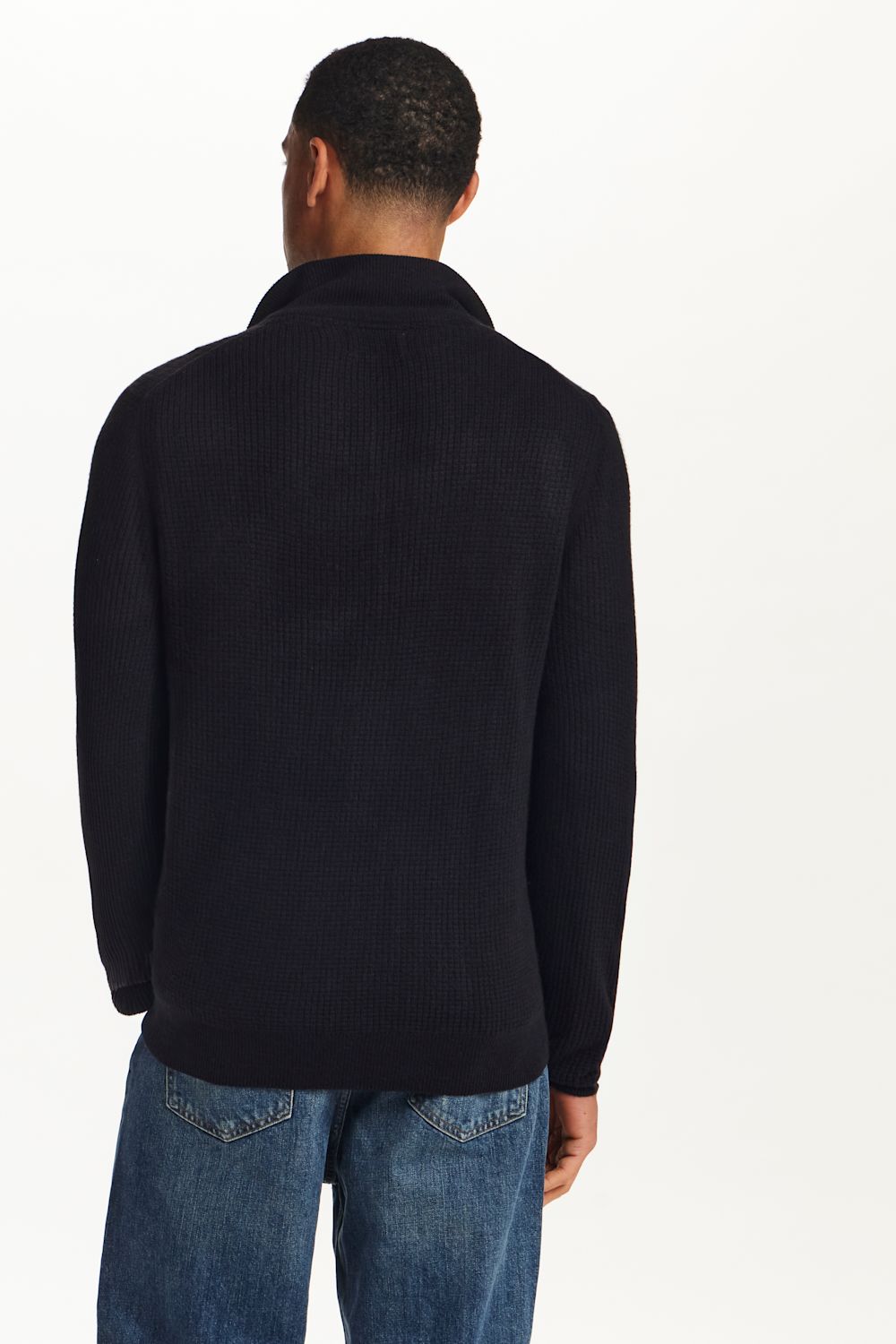 A man viewed from the back wearing a dark navy ribbed knit sweater and blue jeans against a plain white background.