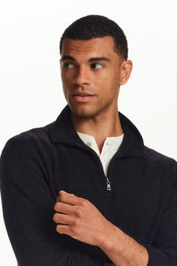 A man wearing a dark navy ribbed quarter-zip sweater over a white collared shirt, photographed against a plain white studio background.
