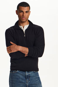 A man wearing a black ribbed quarter-zip mock neck sweater over a white collared shirt and blue jeans, standing with arms crossed against a plain white studio background.