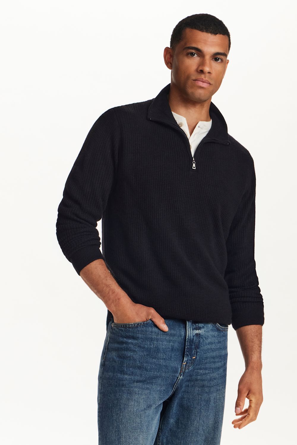 A man wearing a black ribbed quarter-zip mock-neck sweater over a white collared shirt and blue jeans, photographed against a clean white studio background.