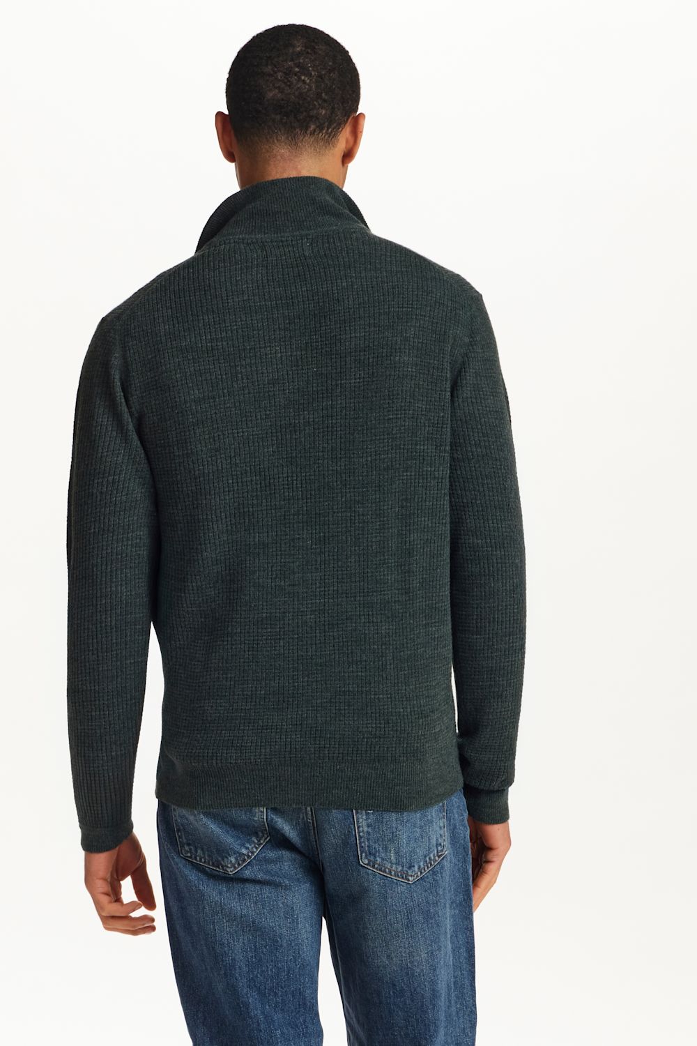 A man viewed from the back wearing a dark green ribbed knit mock neck sweater and blue jeans, against a plain white background.