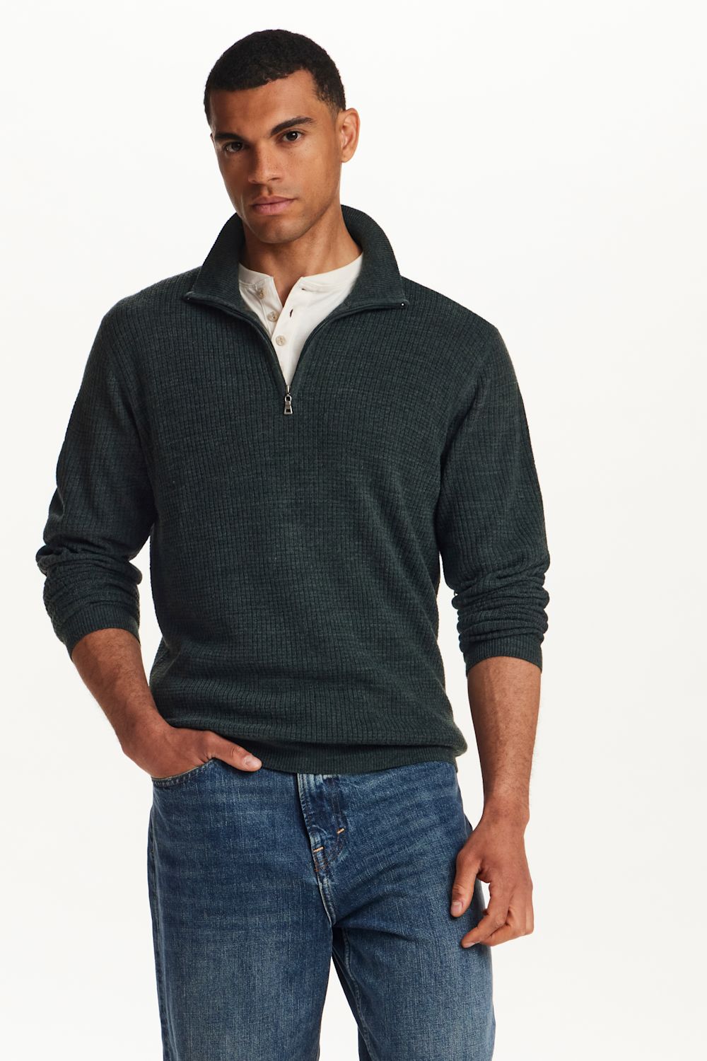 A man wearing a dark green ribbed quarter-zip pullover sweater over a white collared shirt and blue jeans, standing against a plain white studio background.
