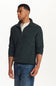 A man wearing a dark green ribbed knit quarter-zip sweater over a white henley shirt and blue jeans, photographed against a plain white studio background.