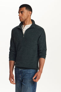 A man wearing a dark green ribbed knit quarter-zip sweater over a white henley shirt and blue jeans, photographed against a plain white studio background.