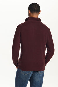 A man viewed from the back wearing a ribbed knit quarter-zip sweater in burgundy and blue jeans, against a plain white background.