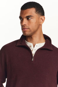 A man wearing a textured waffle-knit quarter-zip pullover sweater in burgundy, layered over a white henley shirt, against a plain white background.