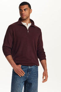 A man wearing a ribbed knit quarter-zip sweater in burgundy, layered over a white collared shirt and paired with blue jeans, photographed against a plain white studio background.