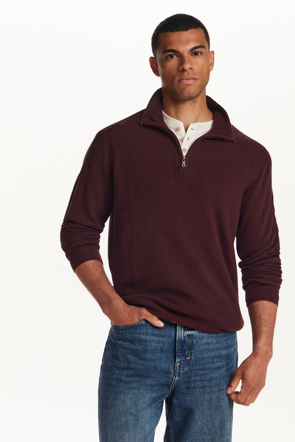 A man wearing a burgundy ribbed quarter-zip sweater over a white collared shirt and blue jeans, standing against a plain white studio background.