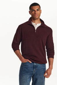 A man wearing a burgundy ribbed quarter-zip sweater over a white collared shirt and blue jeans, standing against a plain white studio background.