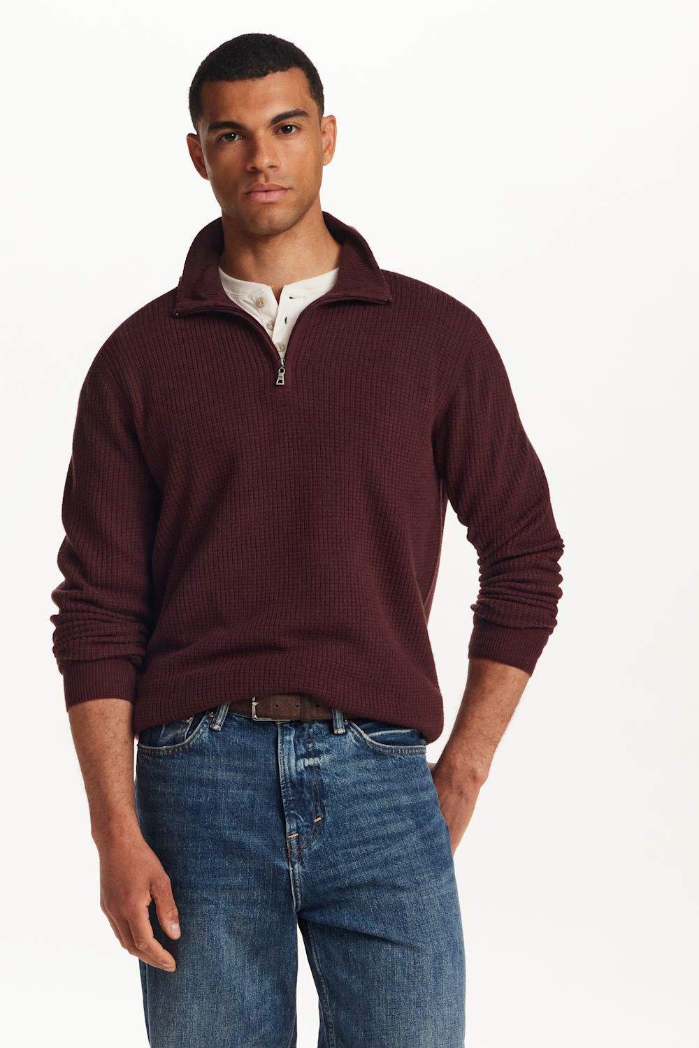 A man wearing a ribbed knit quarter-zip sweater in burgundy, layered over a white collared shirt and paired with blue jeans, photographed against a clean white studio background.