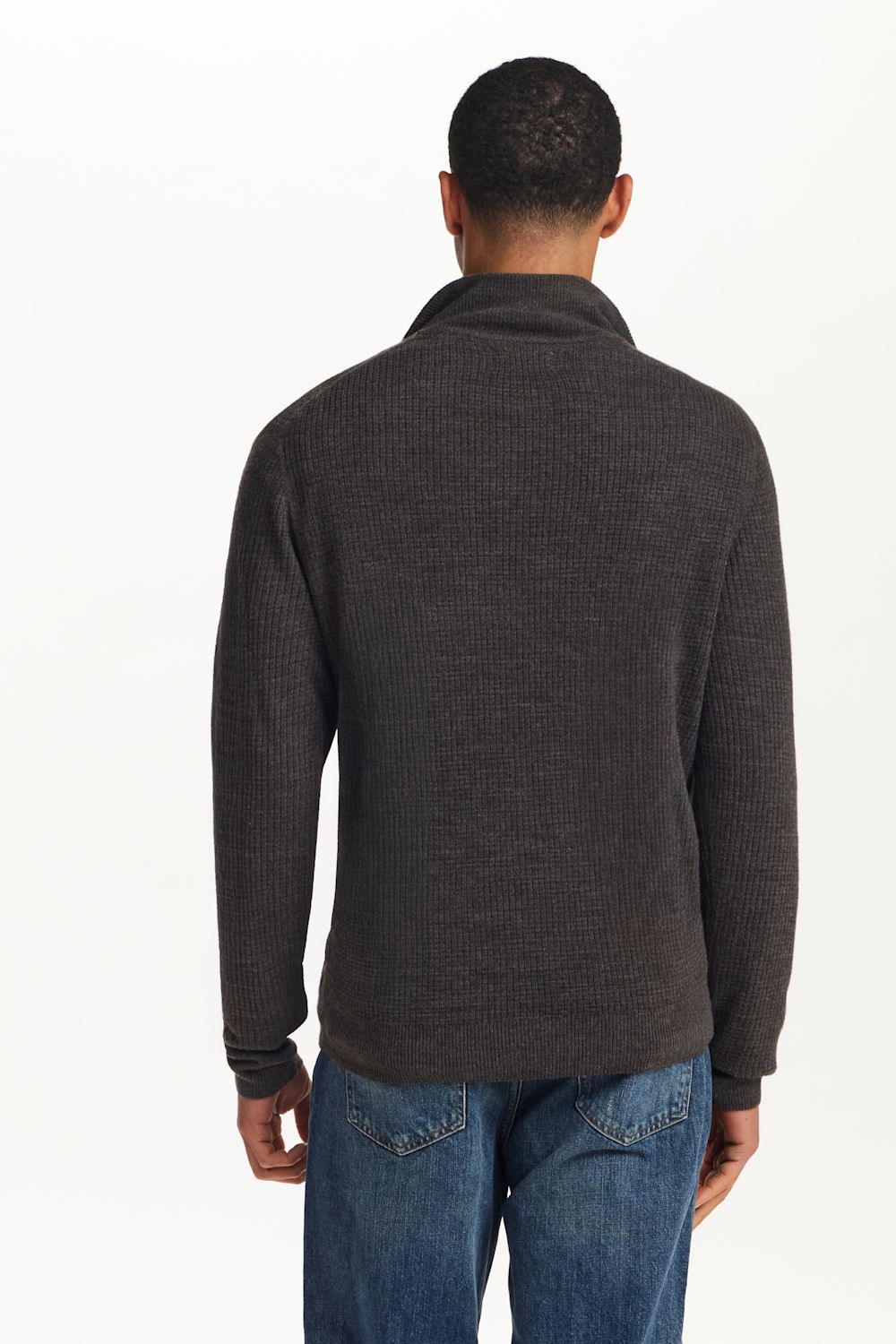 A man viewed from the back wearing a dark charcoal gray ribbed knit mock neck sweater and blue jeans, against a plain white background.