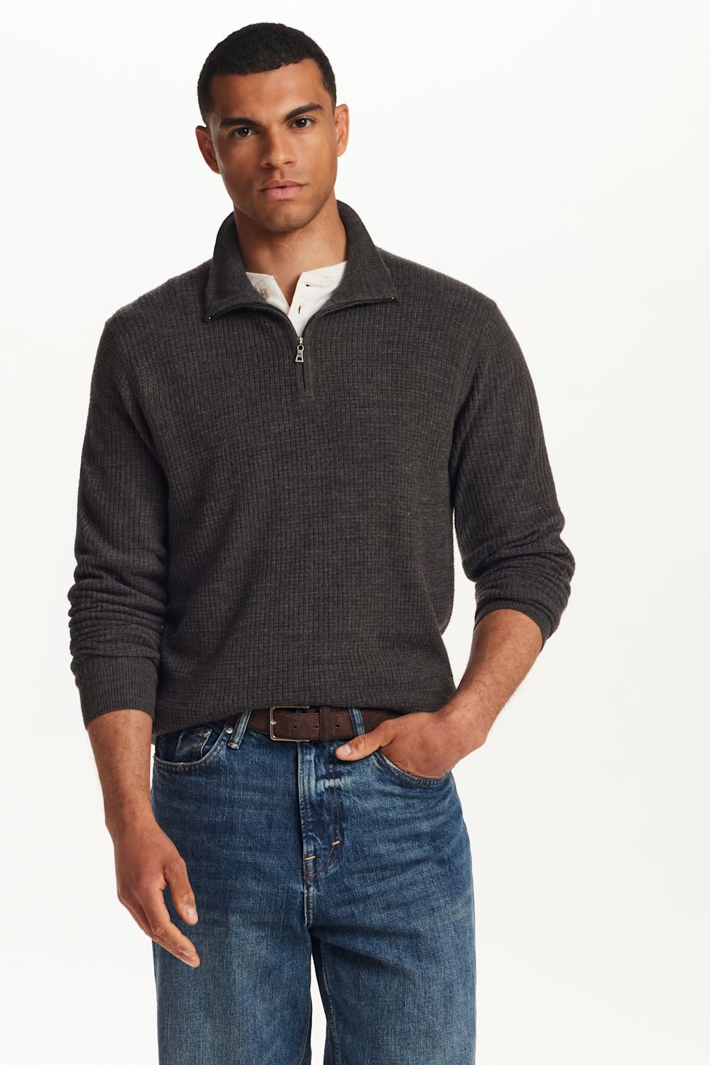 A man wearing a dark charcoal gray ribbed quarter-zip sweater over a white collared shirt and blue jeans, standing against a plain white studio background.