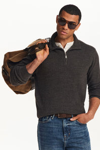 A man wearing a dark charcoal gray ribbed quarter-zip sweater over a white collared shirt, paired with blue jeans and sunglasses, holding a brown duffel bag against a plain white background.