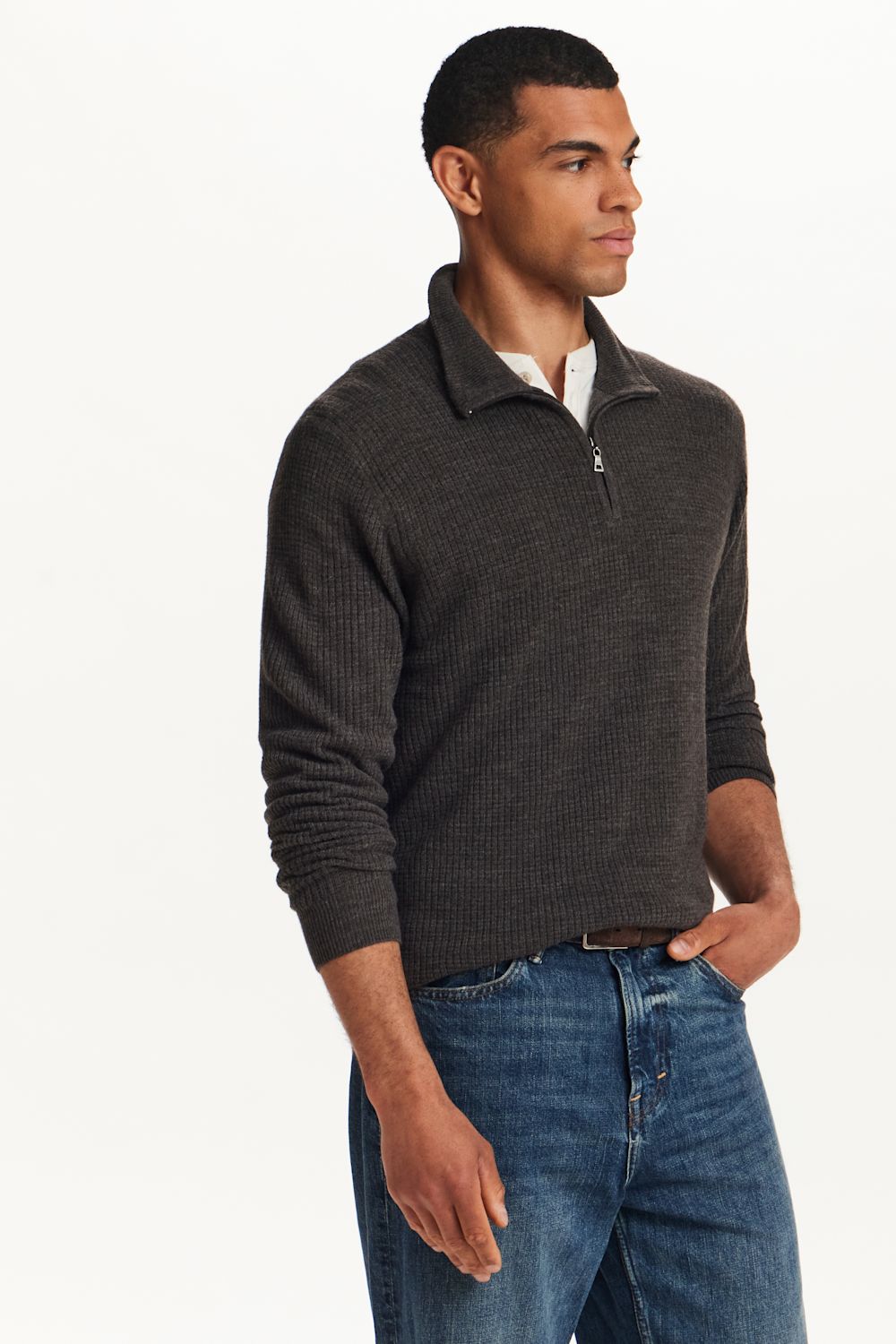 A man wearing a dark charcoal gray ribbed quarter-zip sweater over a white collared shirt and blue jeans, standing against a plain white studio background.