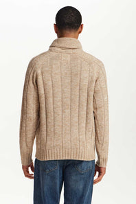 A man viewed from the back wearing a beige ribbed knit turtleneck sweater and blue jeans against a plain white studio background.