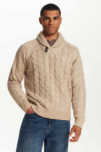 A man wearing a beige cable-knit sweater with a shawl collar, paired with blue jeans, photographed against a plain white studio background.