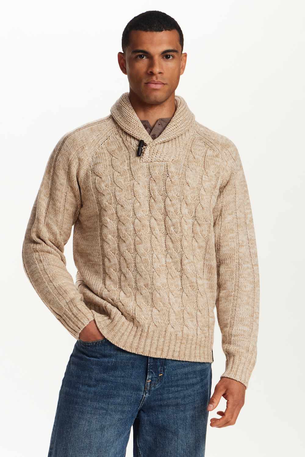 A man wearing a beige cable-knit sweater with a shawl collar, paired with blue jeans, photographed against a plain white studio background.