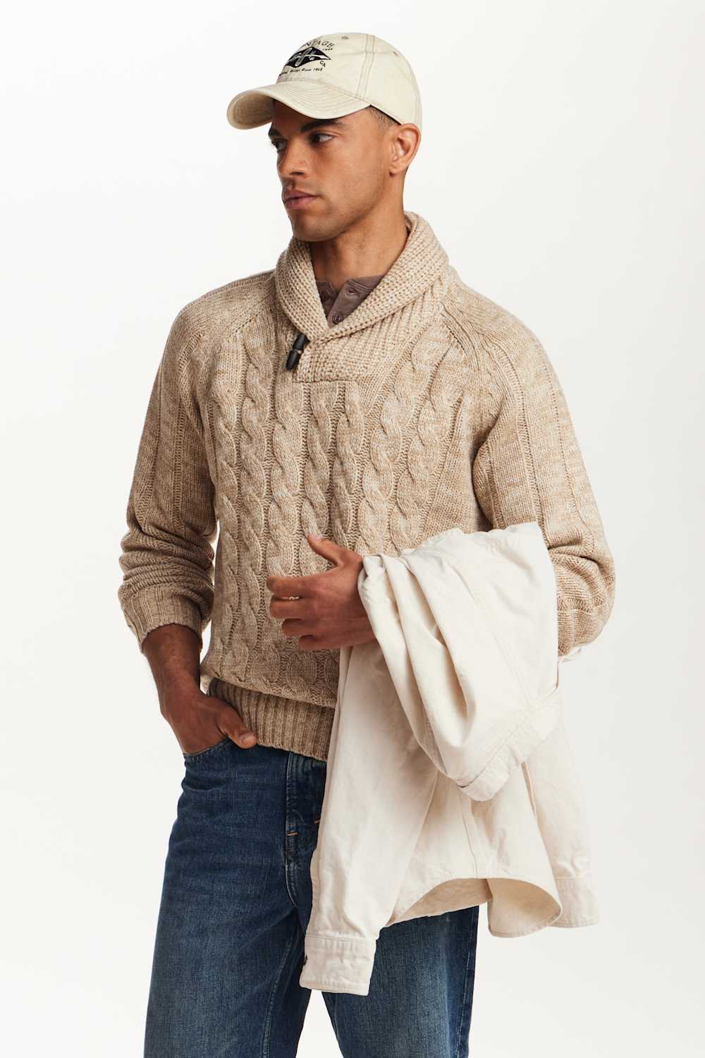 A man wearing a beige cable-knit sweater with a shawl collar, blue jeans, and a cream baseball cap, holding a cream-colored knit sweater over his arm against a plain white studio background.