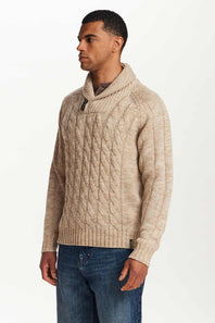 A man wearing a beige cable-knit sweater with a shawl collar, paired with blue jeans, photographed against a plain white studio background.
