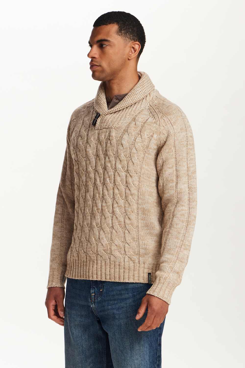 A man wearing a beige cable-knit sweater with a shawl collar, paired with blue jeans, photographed against a plain white studio background.