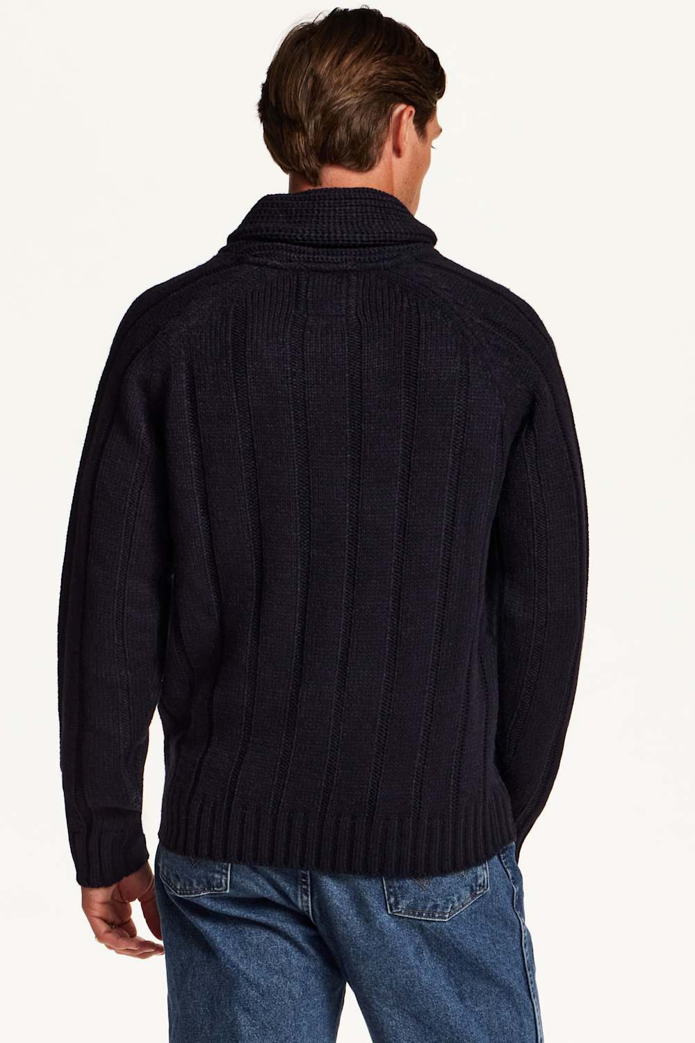 A man viewed from the back wearing a dark navy cable-knit turtleneck sweater and blue jeans against a plain white studio background.