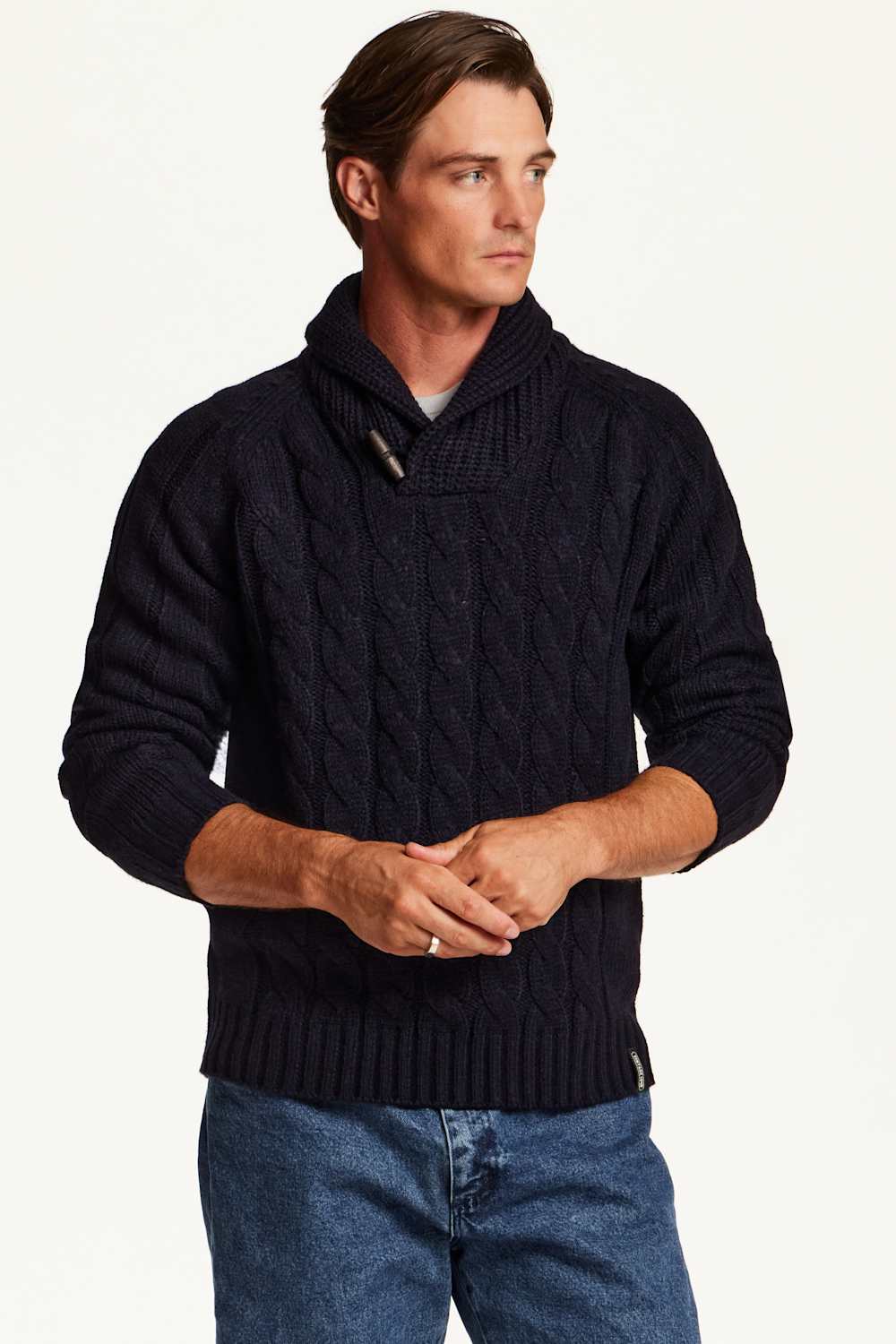 A man wearing a dark navy blue cable-knit sweater with a high folded collar, paired with blue jeans, photographed against a clean white studio background.
