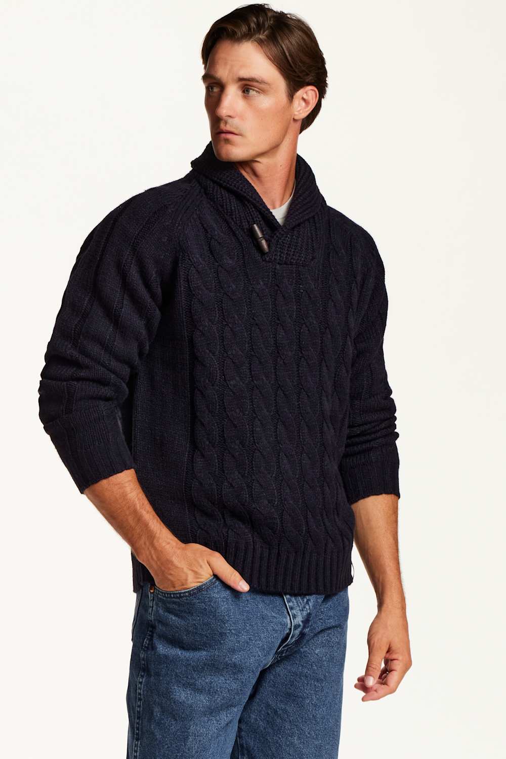 A man wearing a dark navy blue cable-knit sweater with a shawl collar and buttoned placket, paired with blue jeans, photographed against a plain white studio background.