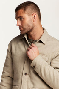 A man wearing a light beige overshirt jacket over a green shirt, shown in a close-up profile view against a plain light background.