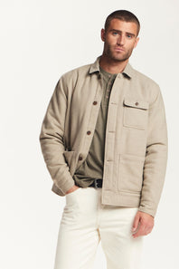 A man wearing a light beige overshirt jacket over an olive green henley shirt and white trousers, standing against a plain white studio background.