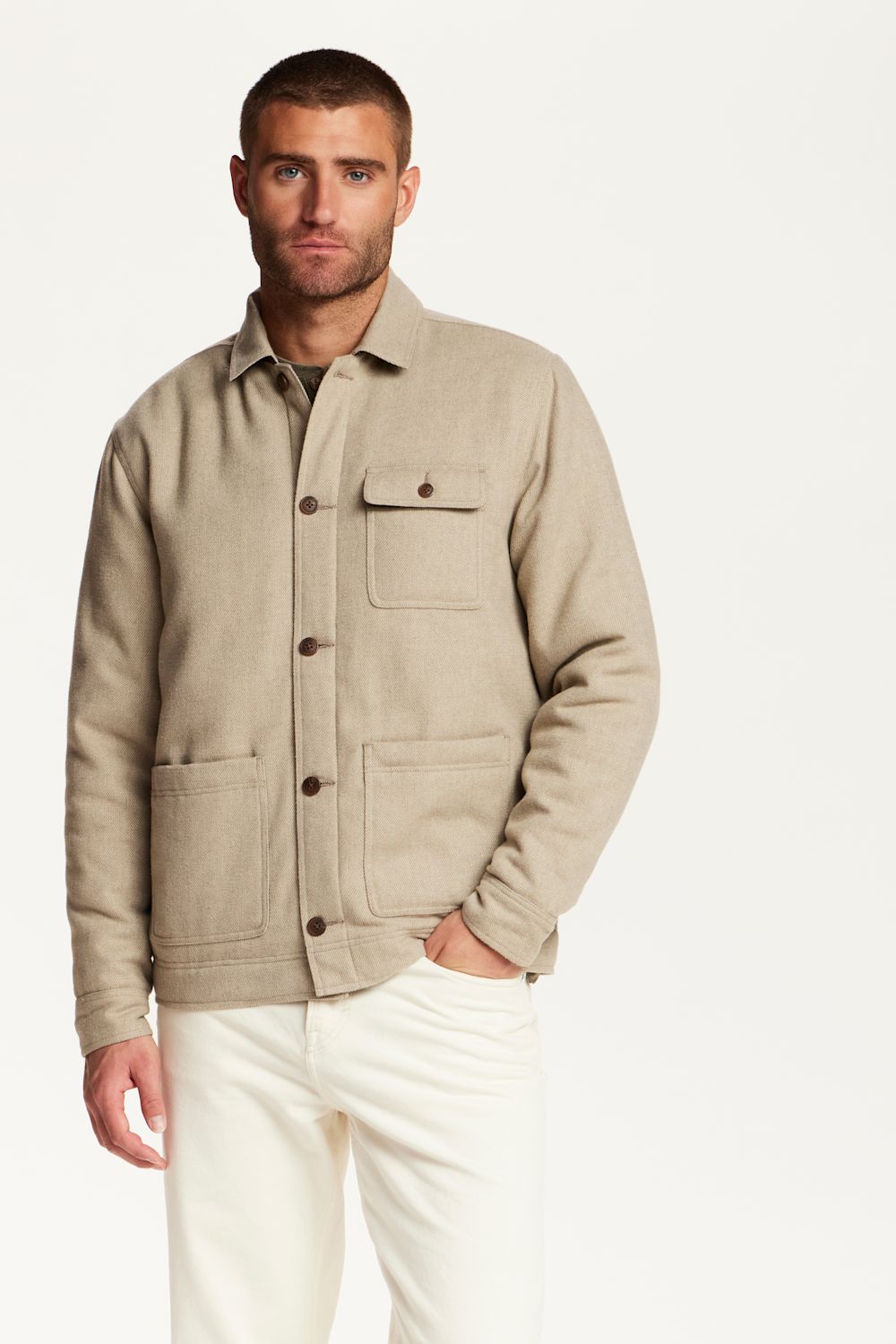 A man wearing a light beige utility jacket with dark buttons and white trousers, standing against a plain white studio background.
