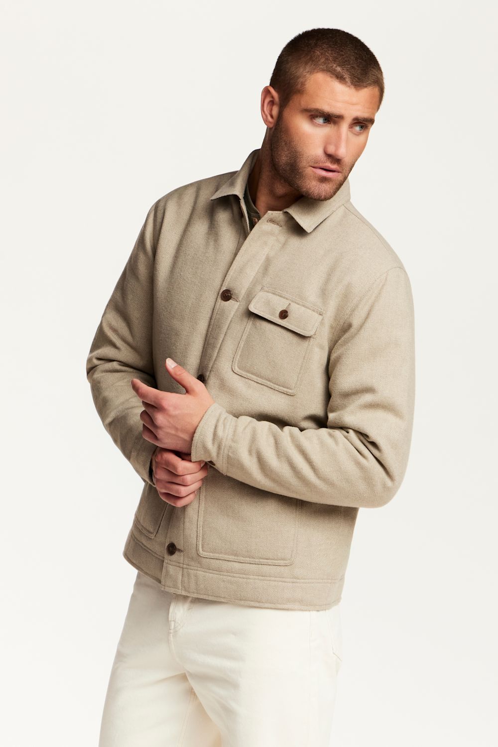 A man wearing a light beige button-up jacket with a chest pocket, paired with off-white trousers, photographed against a clean white studio background.