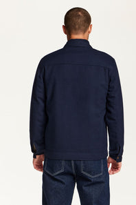 A man viewed from the back wearing a dark navy blue jacket and dark blue jeans against a plain light background.