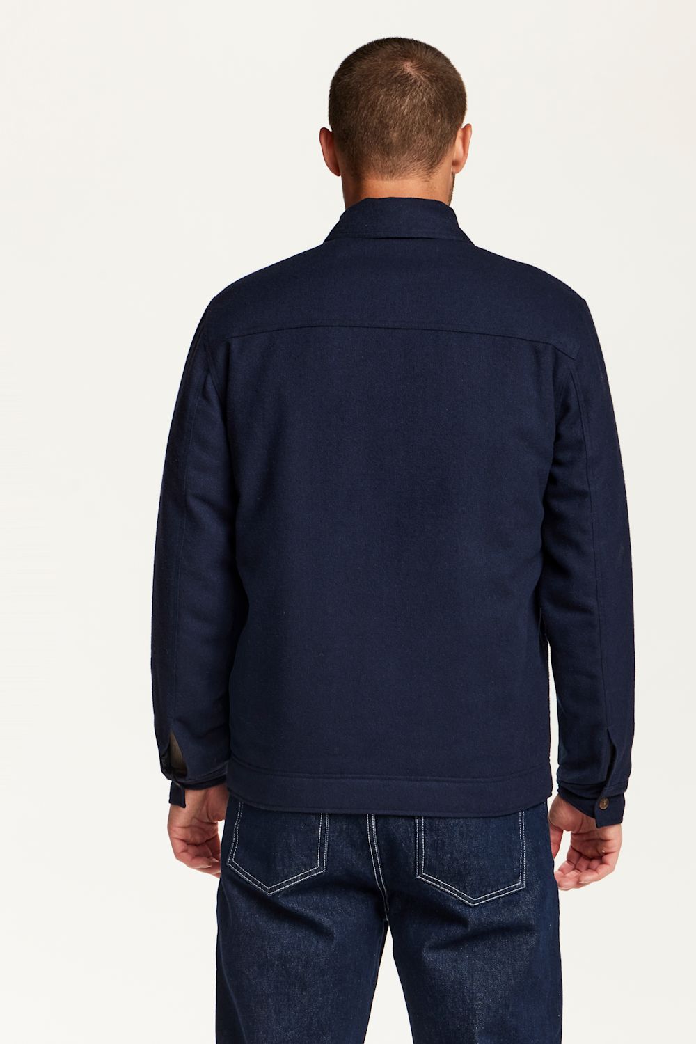A man viewed from the back wearing a dark navy blue jacket and dark blue jeans against a plain light background.