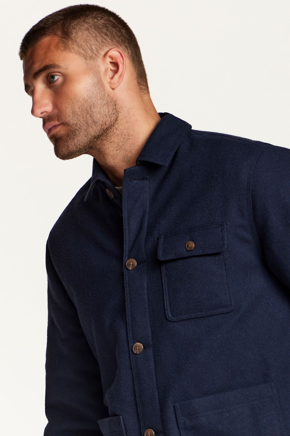 A close-up side profile of a man wearing a dark navy blue overshirt or jacket with a buttoned chest pocket, photographed against a plain light background.