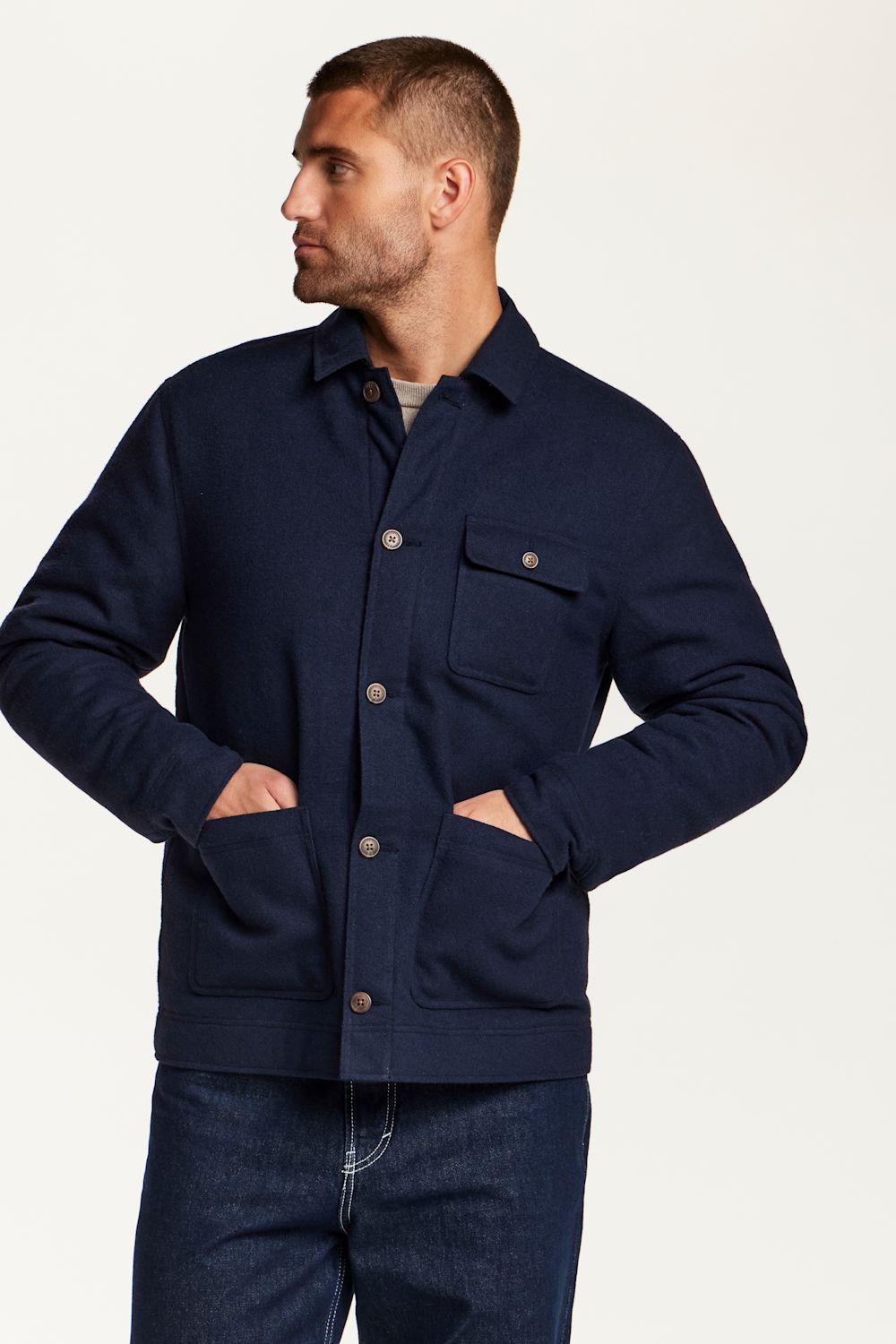 A man wearing a navy blue button-up overshirt jacket with front pockets, paired with dark blue jeans, photographed against a clean off-white studio background.