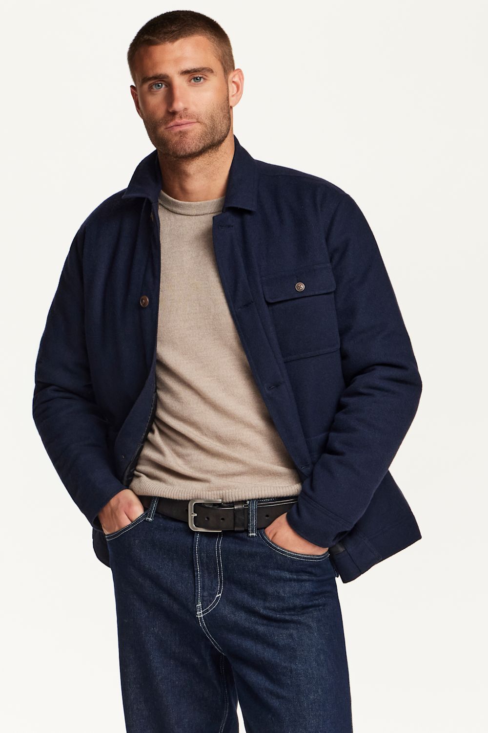 A man wearing a navy blue overshirt jacket over a beige t-shirt and dark blue jeans, standing against a plain off-white background.