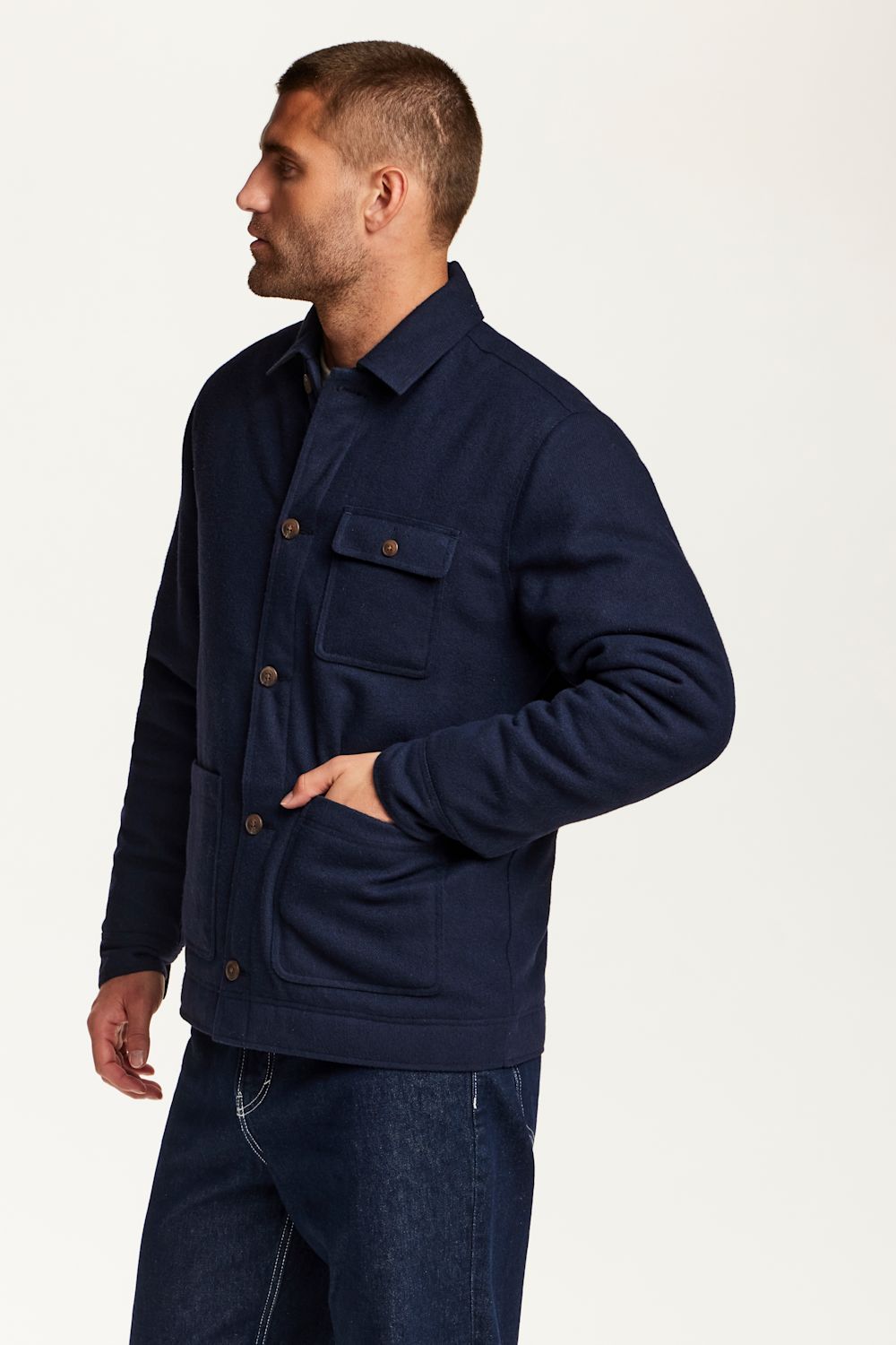 A man wearing a navy blue utility jacket with brown buttons, shown in a side profile against a plain white studio background.