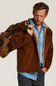 A man wearing a brown suede jacket with a tan sherpa lining over a light blue denim shirt, carrying a brown duffel bag over his shoulder, set against a plain light gray studio background.