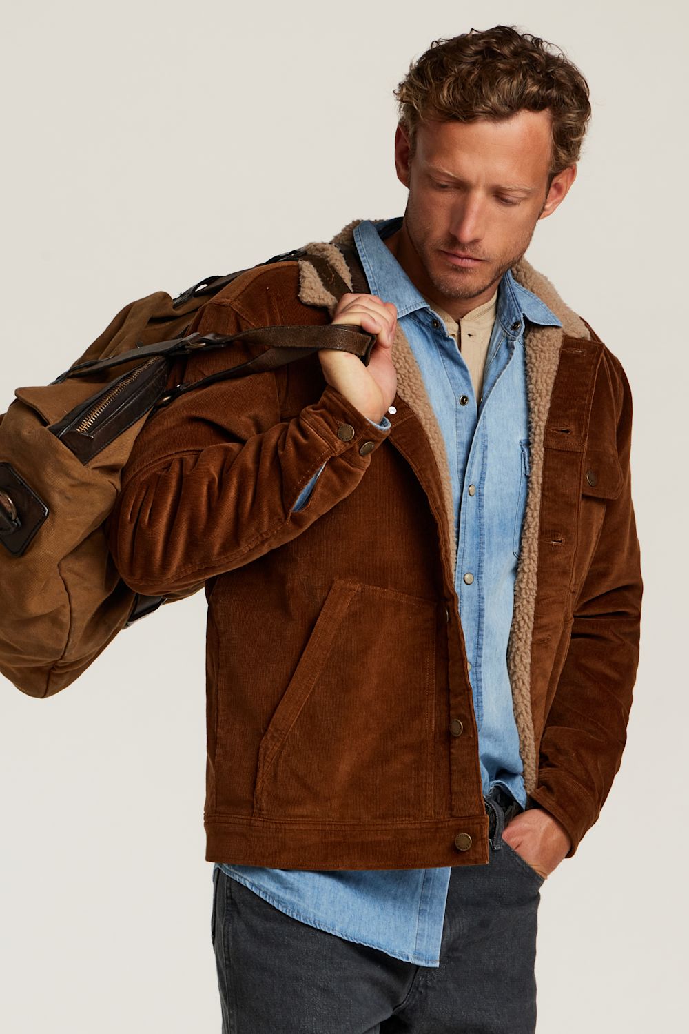A man wearing a brown suede jacket with a tan sherpa lining over a light blue denim shirt, carrying a brown duffel bag over his shoulder, set against a plain light gray studio background.