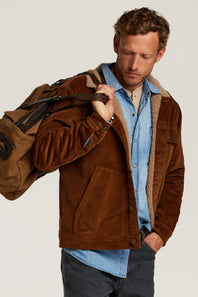 A man wearing a brown suede jacket with a tan sherpa lining over a light blue denim shirt, carrying a brown duffel bag over his shoulder, set against a plain light gray studio background.